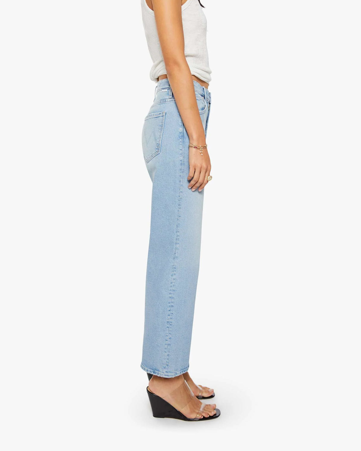 A person wearing The Dodger Ankle jeans by Mother, a sleeveless white top, clear wedge sandals, and gold jewelry stands in profile against a plain white background.