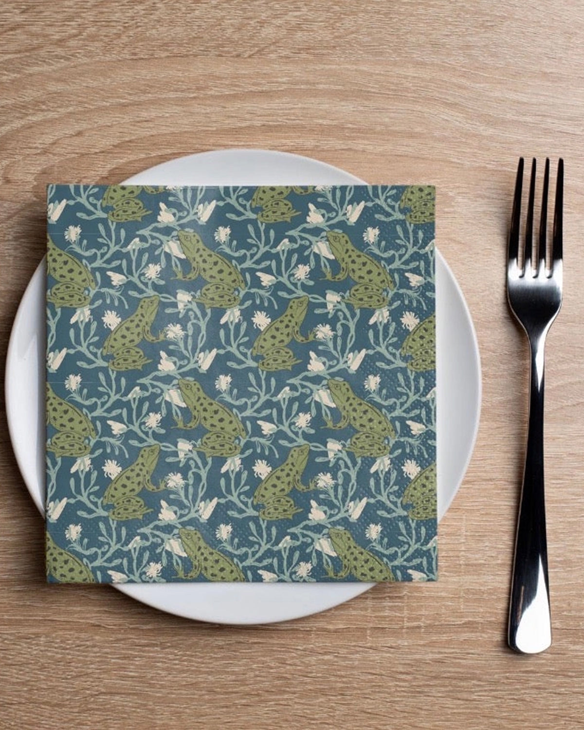 Faire Frog Wallpaper cocktail napkin on a white plate with fork, styled on a wood table by Bungalow Furniture & Accessories.