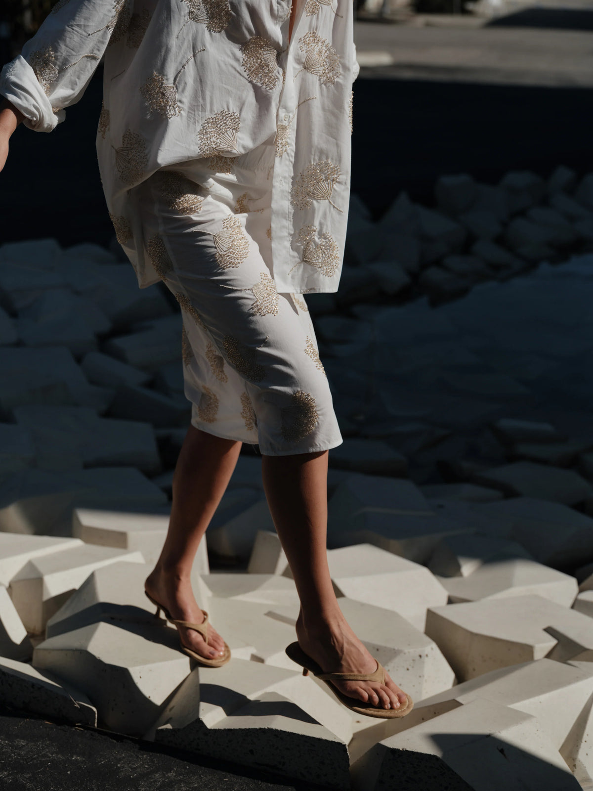 A person in Cissa's The Summer Pencil Skirt walks barefoot on large white geometric blocks, sandals in hand, with dark shadows behind.