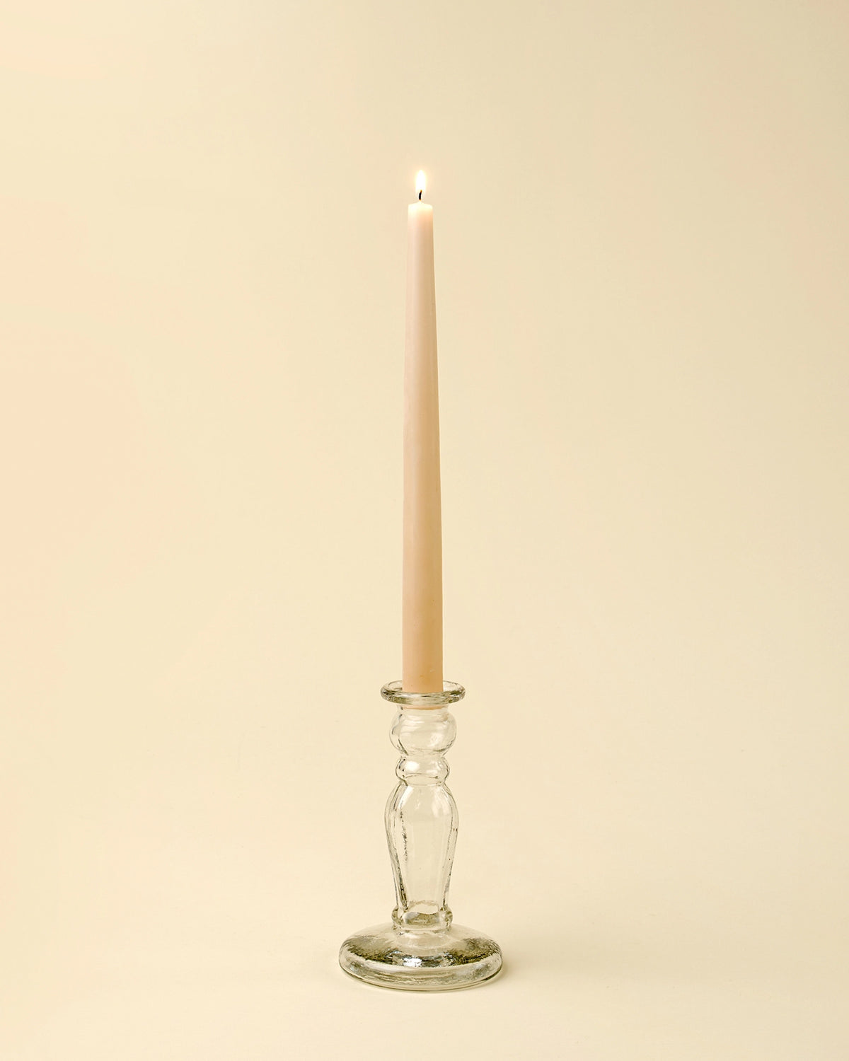 A lit beige taper candle stands upright in the Indaba Vero Glass Candlestick S, crafted from handblown recycled glass, set against a plain, light cream background.