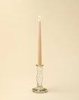 A lit beige taper candle stands upright in the Indaba Vero Glass Candlestick S, crafted from handblown recycled glass, set against a plain, light cream background.