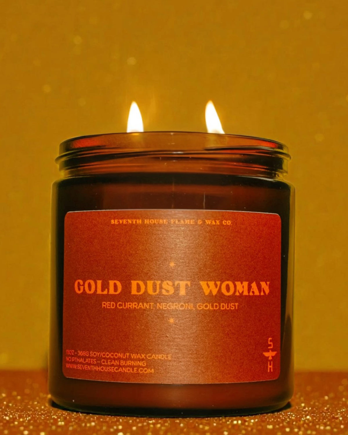 Gold Dust Woman Candle by Faire with two glowing flames, set against a shimmering gold backdrop.
