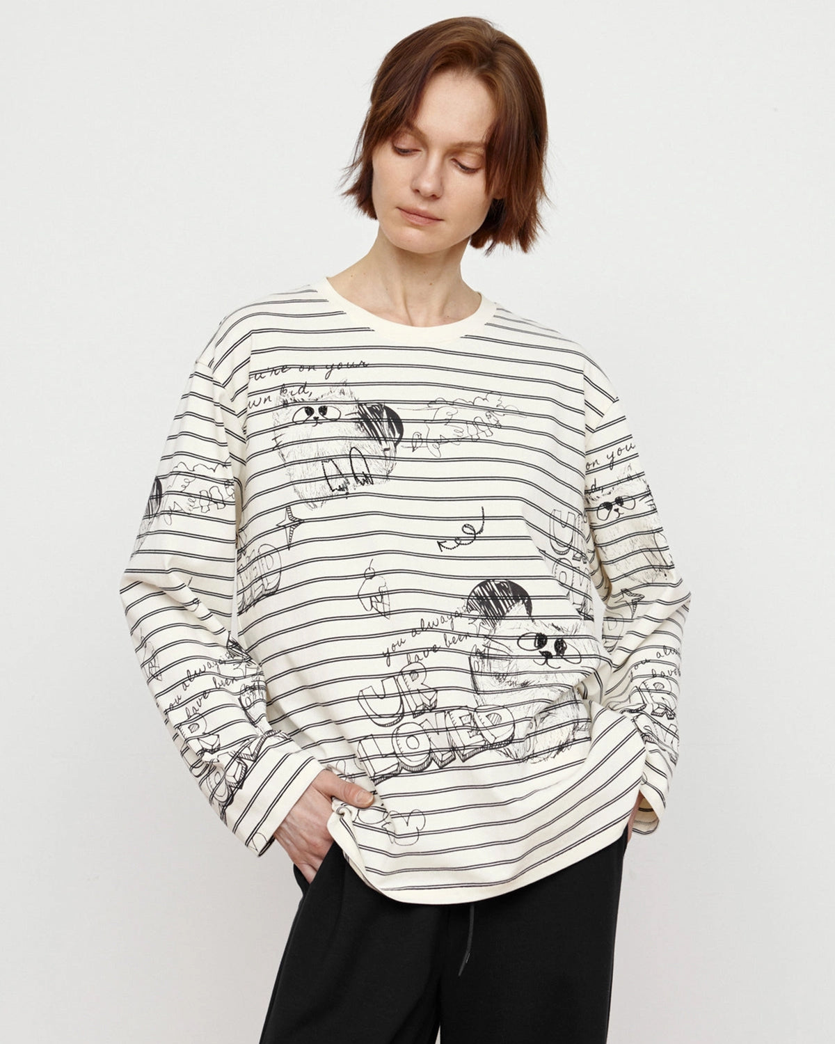 A person with short brown hair wears the Faire Graffiti Stripe Tee, featuring black horizontal stripes and abstract sketches. Looking down while holding the hem, they embody a relaxed street style against a plain background.