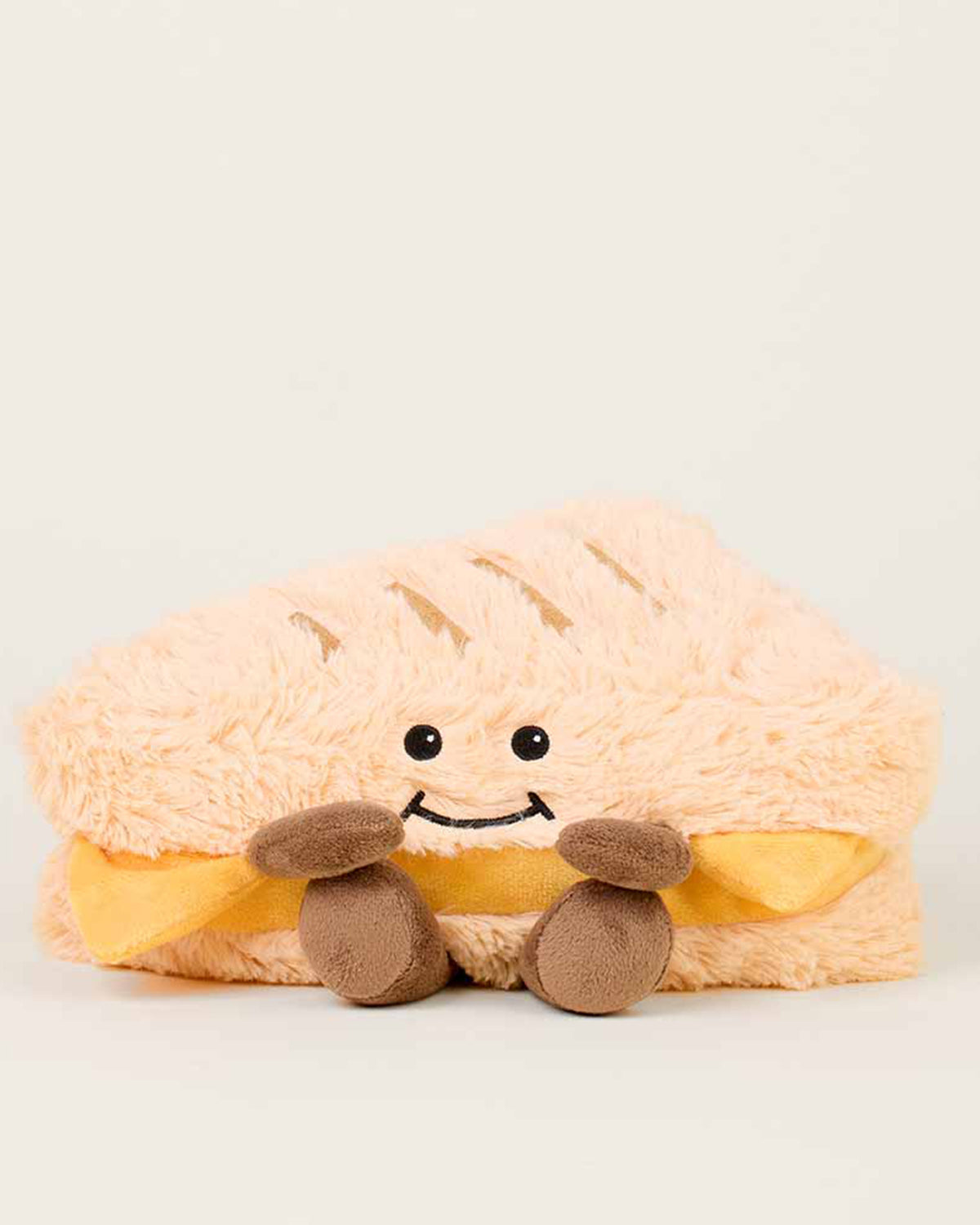 The Faire Grilled Cheese Warmies is a microwavable plush shaped like a smiling grilled cheese, with soft tan "bread," yellow "cheese," brown plush hands and feet, black eyes, and a cheerful smile, providing soothing warmth.