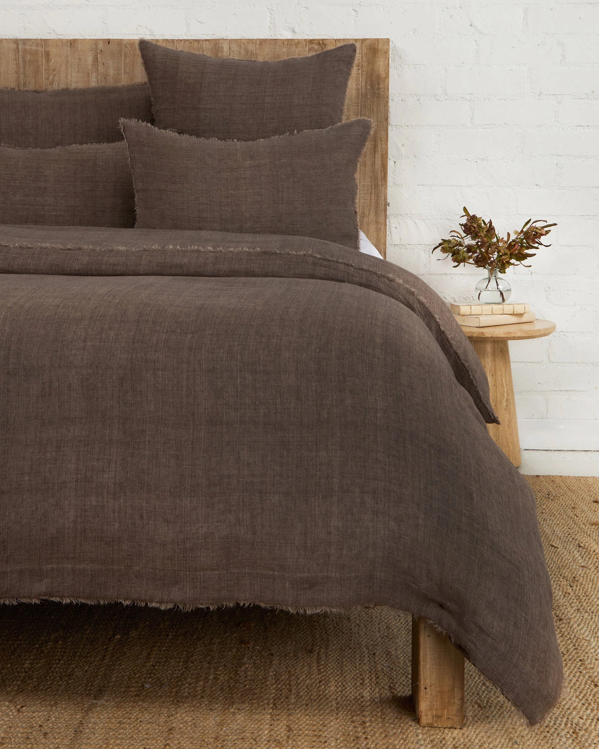 A rustic bed with a wooden headboard features the Hunter Duvet by POM POM and brown pillows. A small wooden bedside table holds a floral vase. The room includes a white brick wall and a woven rug.