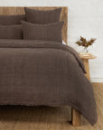 A rustic bed with a wooden headboard features the Hunter Duvet by POM POM and brown pillows. A small wooden bedside table holds a floral vase. The room includes a white brick wall and a woven rug.