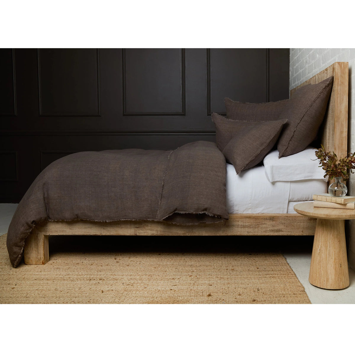 A wooden bed topped with the Hunter Duvet and linen shams from POM POM rests against a dark-paneled wall. Nearby, a round wooden table holds books and a vase of flowers on a woven rug.