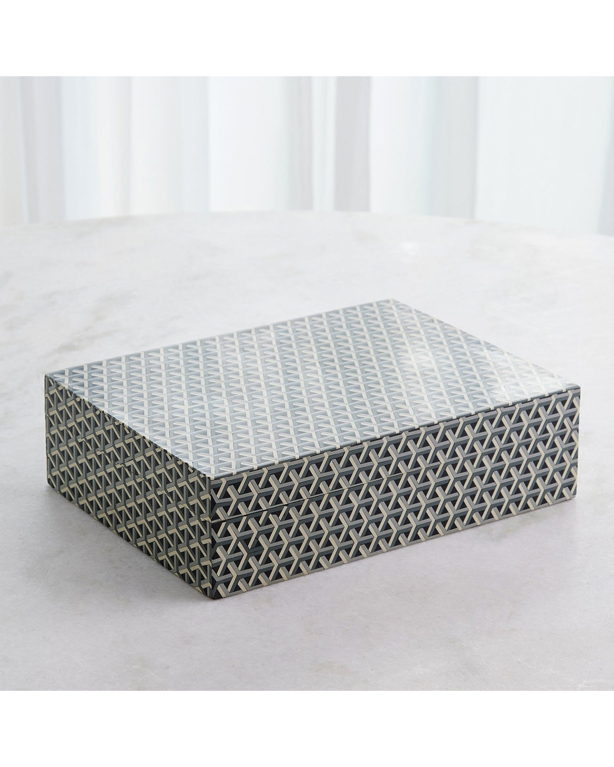 The Interwoven Box LG by Global Views features a rectangular shape with a grey and white geometric pattern, ideal for decorative storage. Shown on a light surface with white curtains in the background.