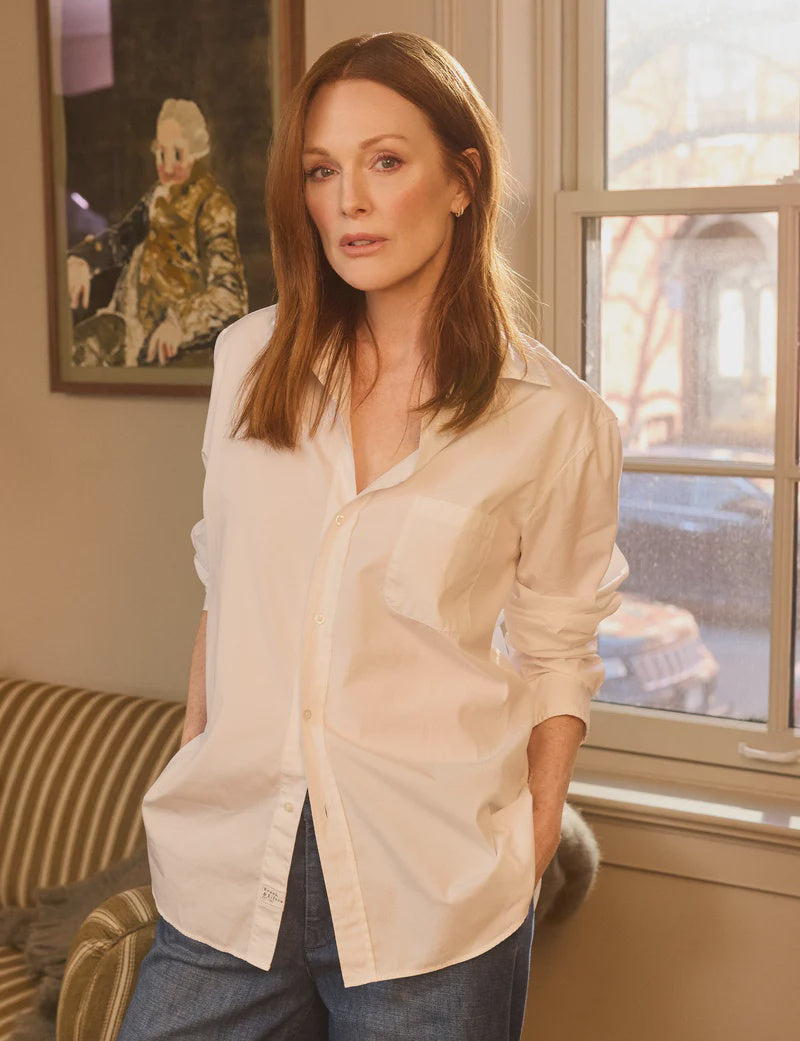 A red-haired woman channeling Julianne Moore stands in a room, wearing a WAVERLY Relaxed Button-Up Shirt by Frank & Eileen and tailored-fit jeans. She gazes at the camera neutrally as a window behind her frames a painting on the wall.