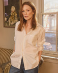 A red-haired woman channeling Julianne Moore stands in a room, wearing a WAVERLY Relaxed Button-Up Shirt by Frank & Eileen and tailored-fit jeans. She gazes at the camera neutrally as a window behind her frames a painting on the wall.