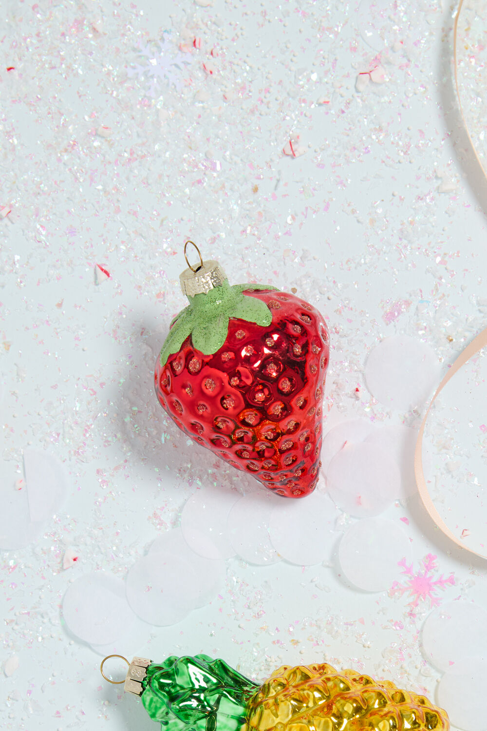 The Juicy Ornament by Accent Decor, a glass strawberry-shaped Christmas ornament (2.25" x 2.25" x 3") with a green top, rests on a sparkly surface dusted with white confetti and a light pink ribbon nearby.