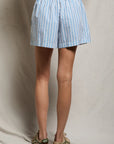 A person’s lower half is shown wearing the Perfectwhitetee Julie Poplin Short—light blue and white vertical stripes with a smocked elastic waistband—paired with tan sandals, set against a neutral fabric backdrop.