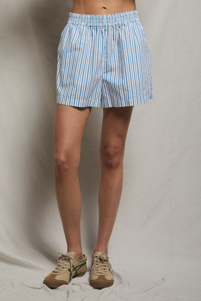 Wearing the Julie Poplin Short by Perfectwhitetee—light blue and white striped cotton with a smocked waist—paired with beige sneakers with green accents, standing against a plain light-colored background for a true high-summer staple look.