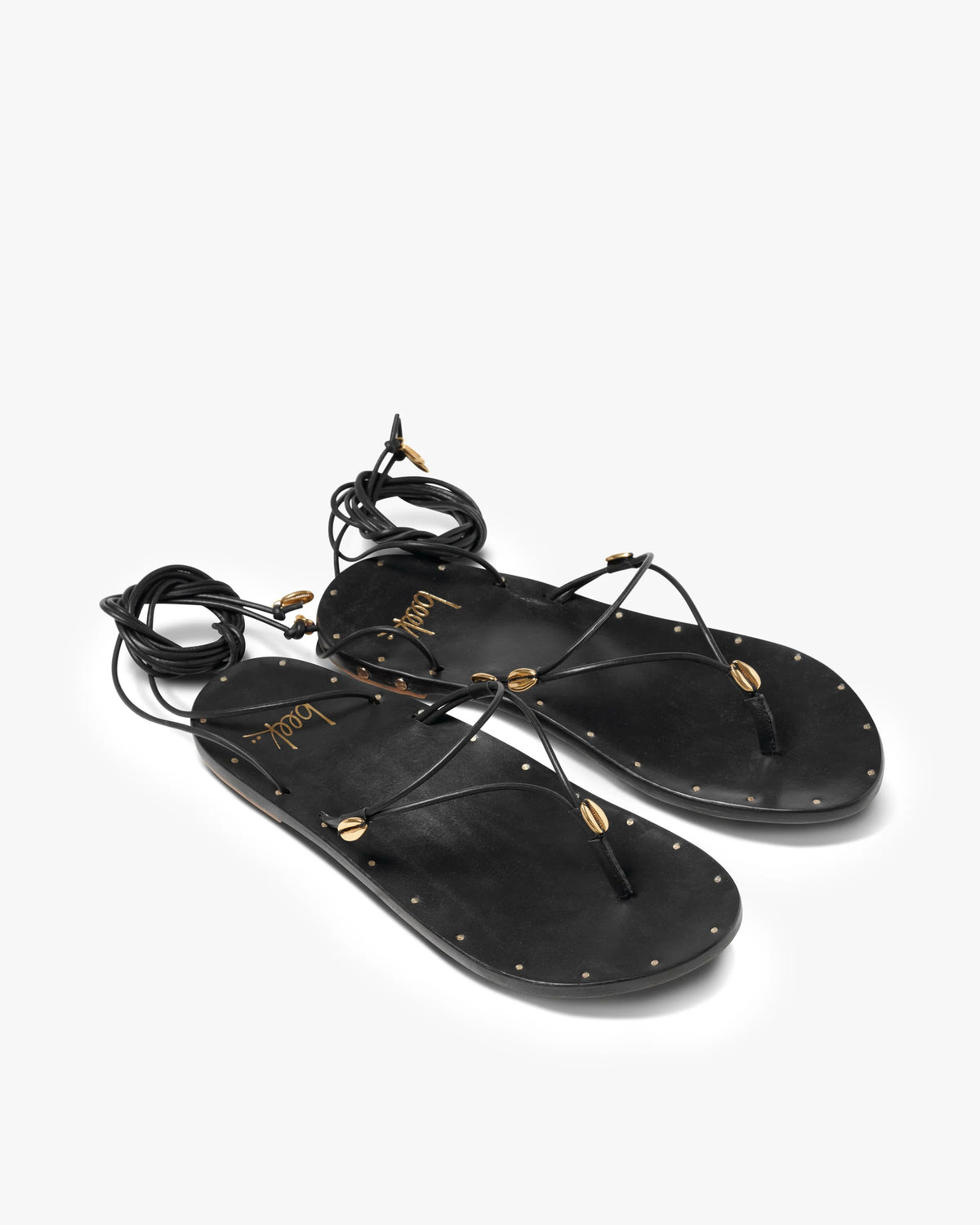 The Beek Koha Sandal features black leather, adjustable ankle-tie lace-up straps, gold bead and shell accents, and small gold studs outlining the soles, shown on a white background.