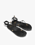 The Beek Koha Sandal features black leather, adjustable ankle-tie lace-up straps, gold bead and shell accents, and small gold studs outlining the soles, shown on a white background.
