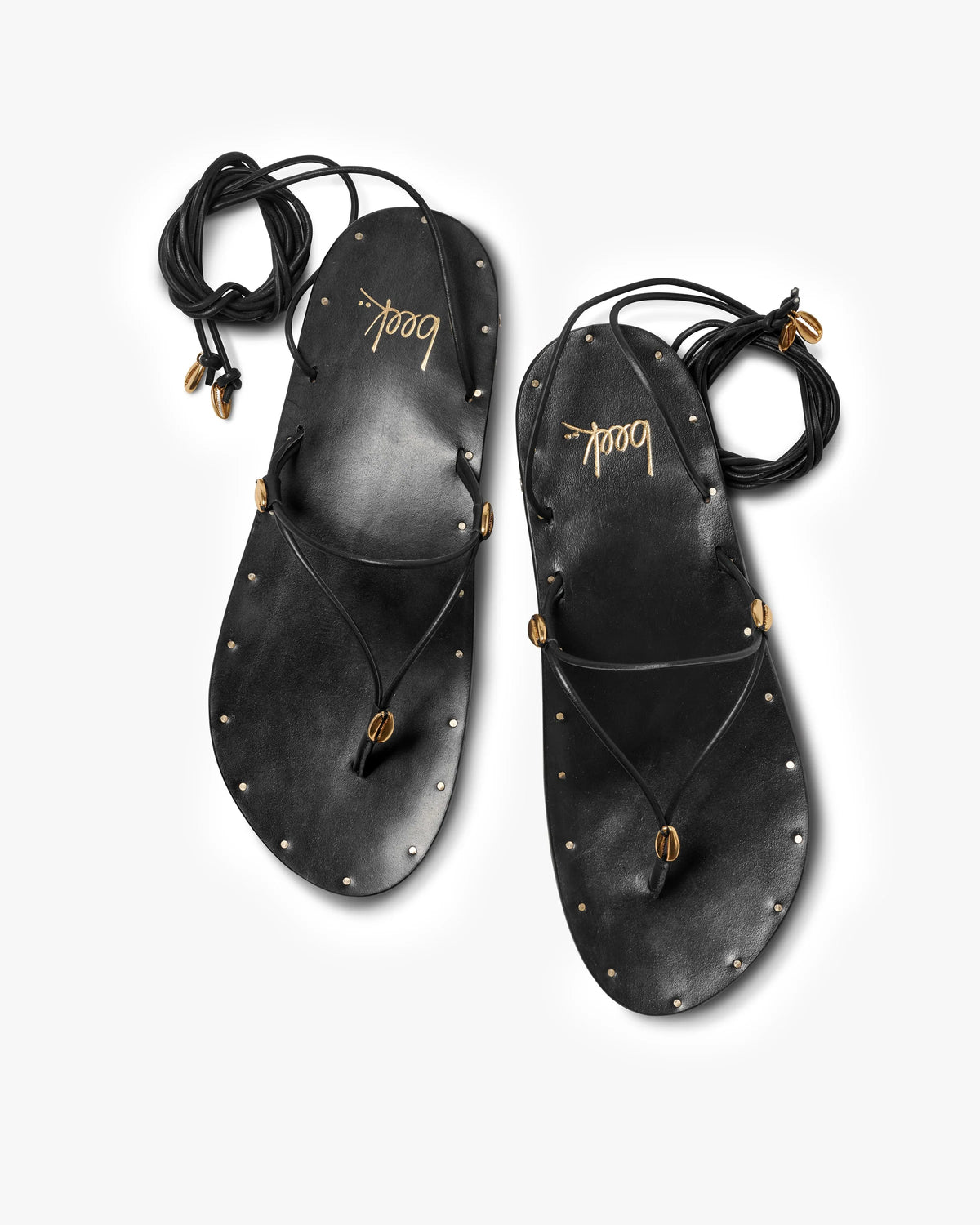 A pair of black leather Koha Sandals by Beek, featuring adjustable ankle-tie laces, gold bead accents, and metal studs around the edges, displayed on a white background. "Beek" is printed in gold on each insole.