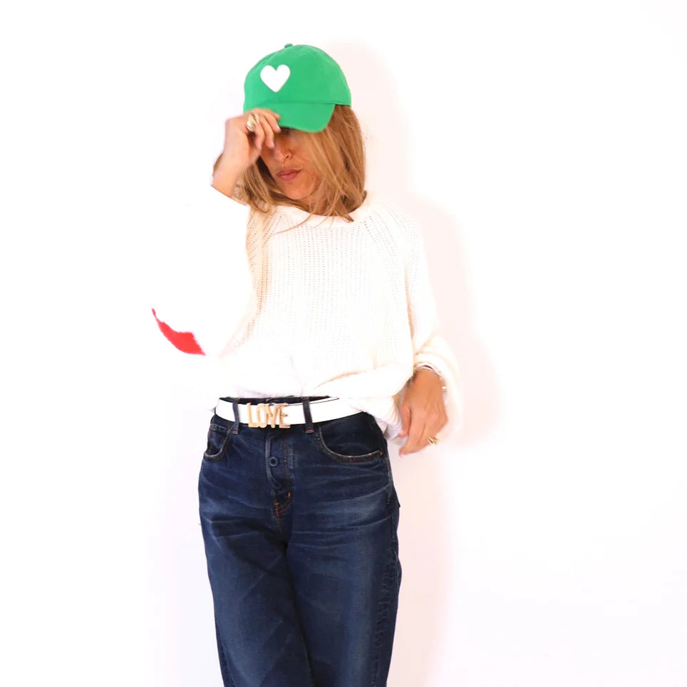 A woman in a white sweater and dark jeans adjusts her KR Imperfect Heart Hat from Kerri Rosenthal, featuring its signature exclusive color and white heart. Her "LOVE" belt accentuates her effortless chic pose against the plain white backdrop, embodying a timeless KR staple.