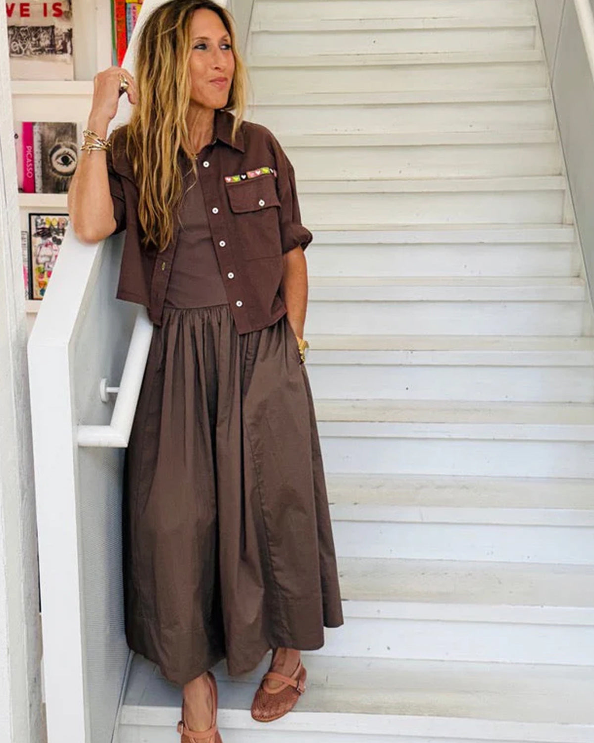 A woman stands on white stairs wearing the KR Tank Dress by Kerri Rosenthal with a matching short-sleeved jacket. She has long hair, sandals, bracelets, and colorful books and art appear in the background.