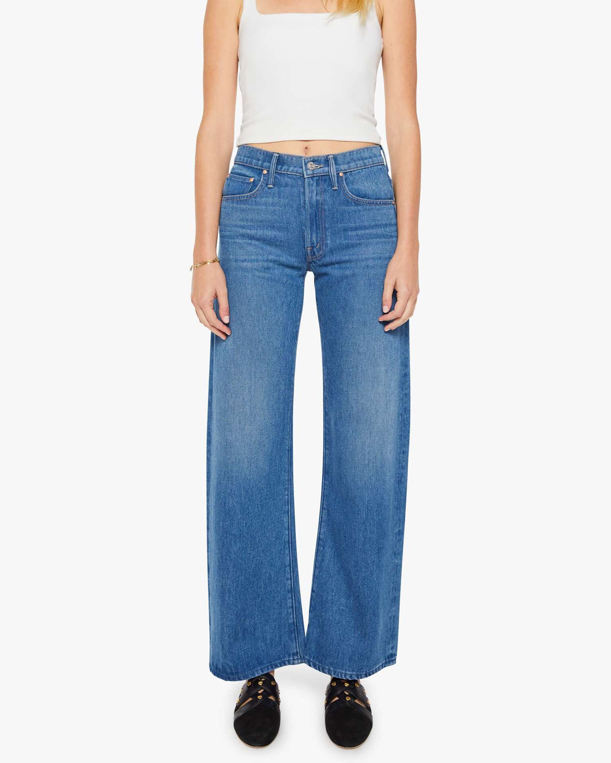 A woman stands facing forward against a white background, wearing Mother’s The Mid Rise Lasso Sneak jeans with a white sleeveless crop top and black shoes.