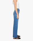 A woman stands in profile against a plain white background wearing a white sleeveless top, Mother’s The Mid Rise Lasso Sneak denim wide-leg pants, and black flat strap shoes.