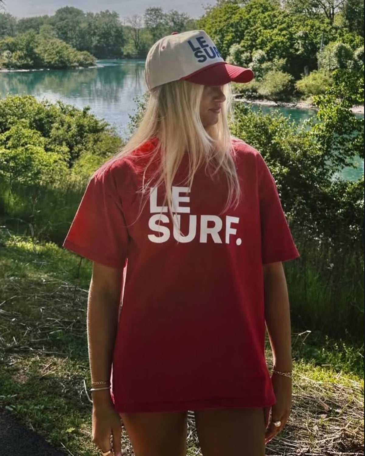 A person with long blonde hair wears a Ping Pong Surf Club Le Surf Tee and stands by a lake under a sunny sky, green trees in the background, with an impressive surfboard beside them.