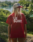 A person with long blonde hair wears a Ping Pong Surf Club Le Surf Tee and stands by a lake under a sunny sky, green trees in the background, with an impressive surfboard beside them.