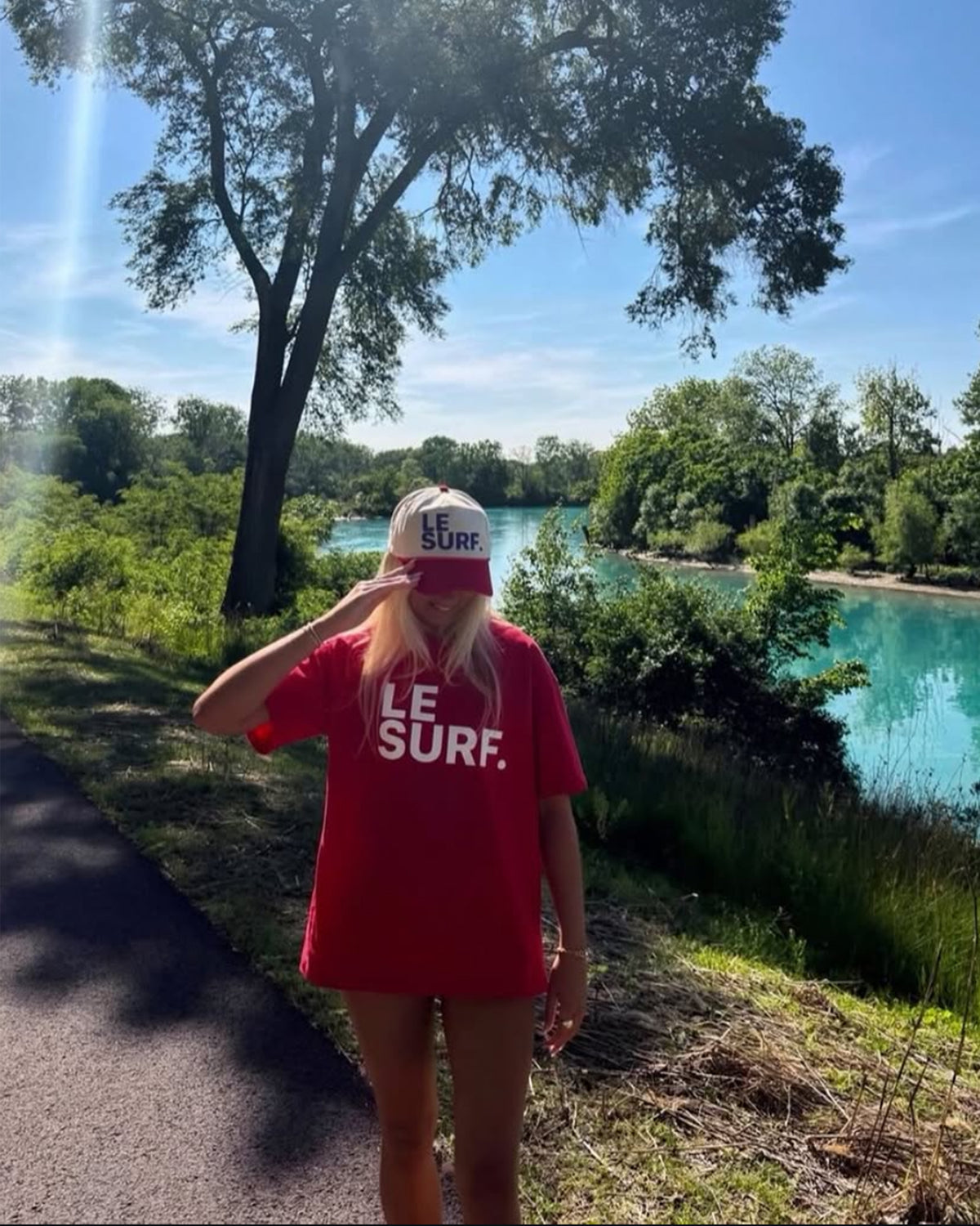 A person in the Le Surf Tee by Ping Pong Surf Club and a matching cap stands on a riverside path, shaded by trees, with a classic surfboard nearby and a vivid blue river framed by lush greenery in the background.