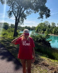 A person in the Le Surf Tee by Ping Pong Surf Club and a matching cap stands on a riverside path, shaded by trees, with a classic surfboard nearby and a vivid blue river framed by lush greenery in the background.