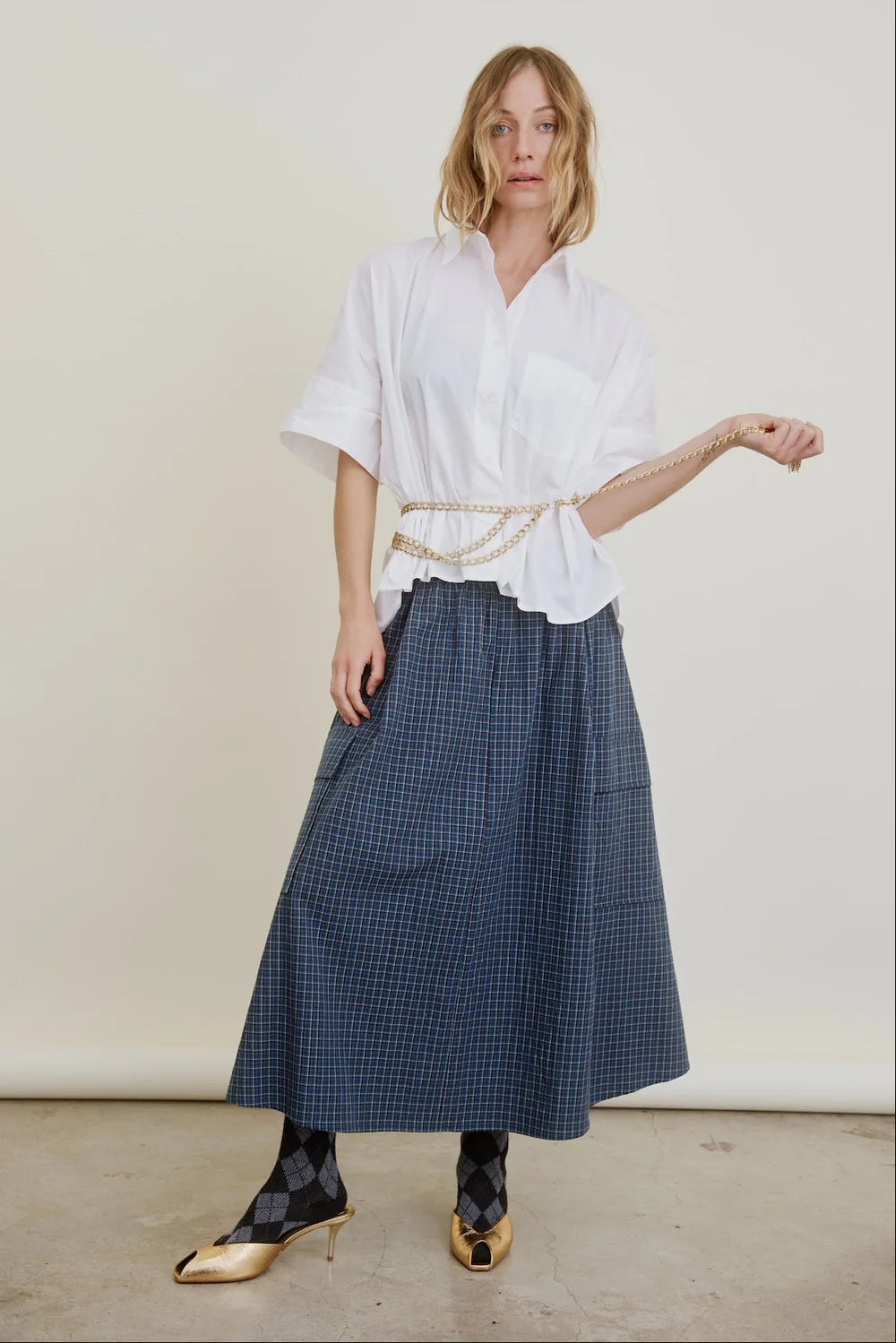 A woman poses against a plain background in a loose white button-up shirt, Aquarius Cocktail Le Cargo Skirt in blue plaid Japanese cotton poplin, argyle socks, gold pointed heels, and a chain belt at her waist.