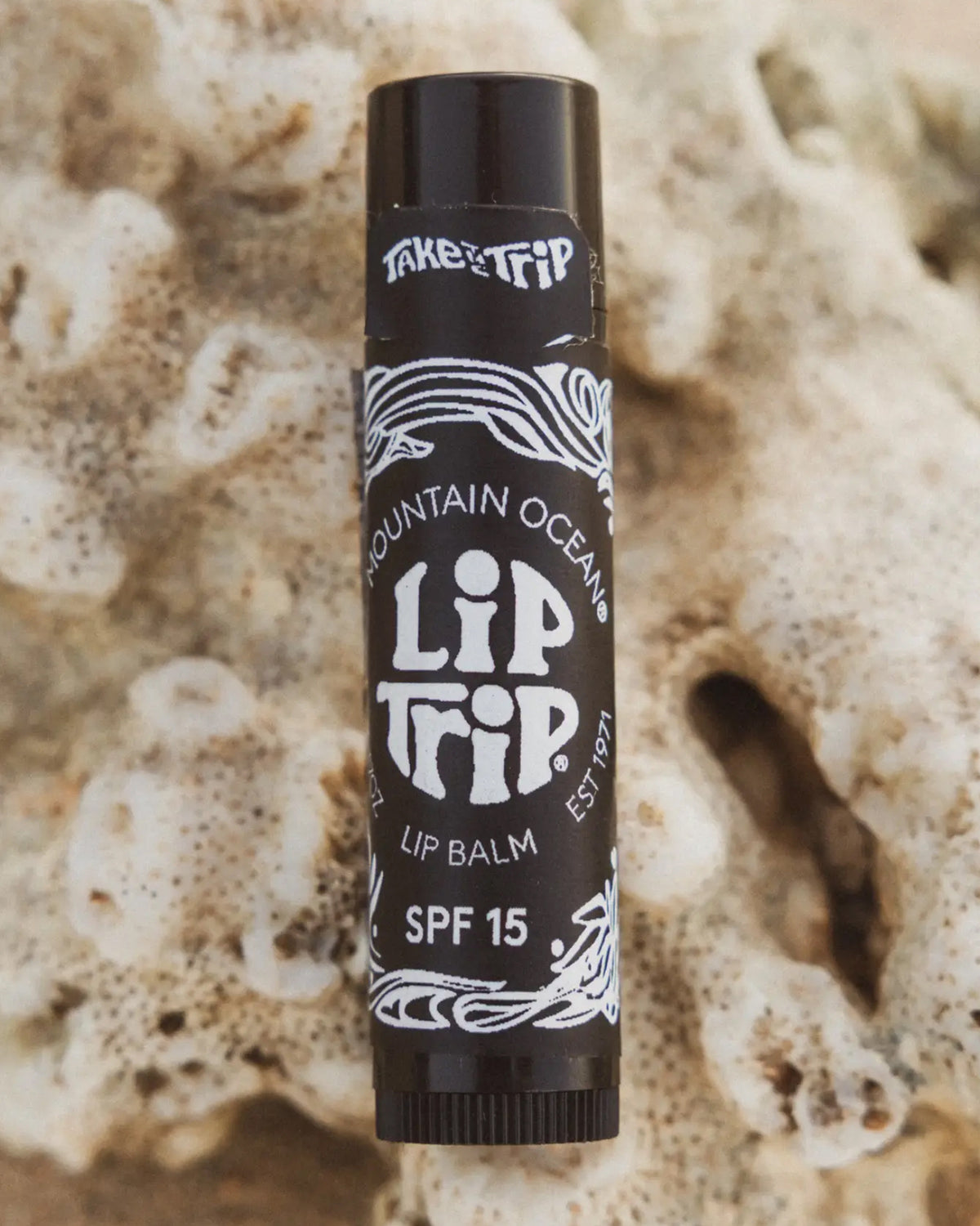 A black tube of Faire Lip Trip SPF lip balm sits on a textured beige surface resembling coral, with white decorative accents and bold text on the label emphasizing its sun protection benefits.