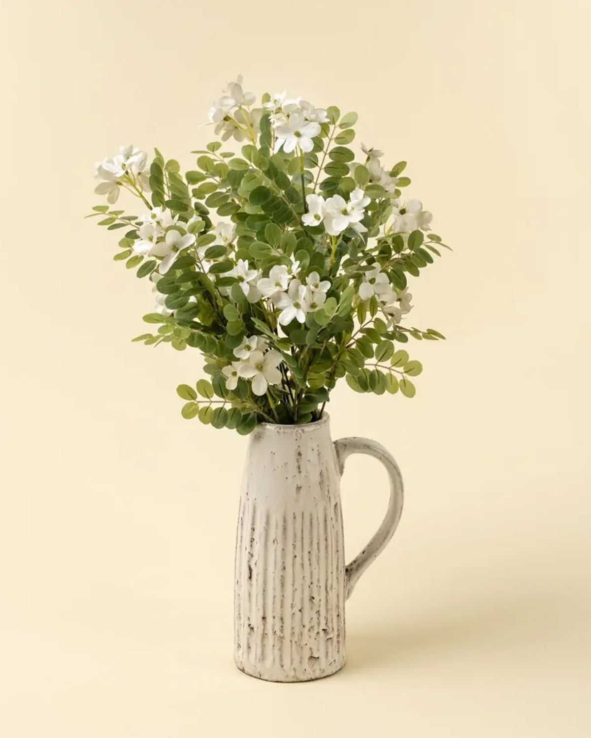 A white ceramic pitcher holds green foliage and Indaba's 01-9528 Lotus Flower Branches, creating elegant accent decor against a plain beige background.