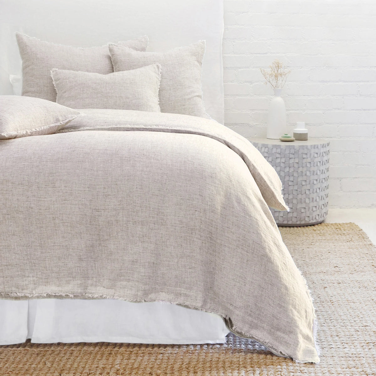 In a cozy bedroom scene, a neatly made bed is adorned with beige linen bedding and matching pillows from the Logan Duvet by POM POM. A textured rug covers the floor, and a round side table displays a vase and candle, all set against a white brick wall for that perfect heathered effect.