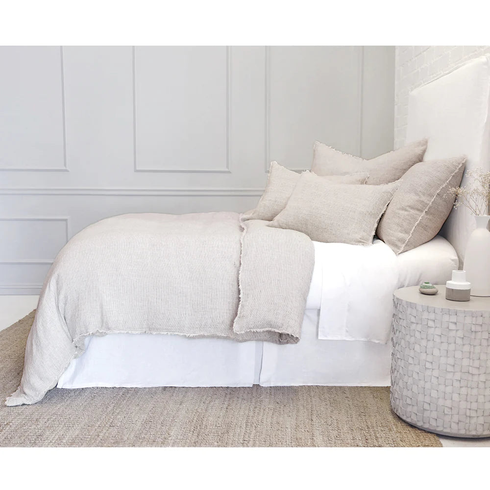 The minimalist bedroom features a neatly made bed adorned with POM POM's Logan Duvet in light beige linen, complemented by pillows and a textured bedspread. Next to the bed is a round, woven side table set against walls with subtle panel detailing. The floor showcases a natural woven rug from the Loga Collection, offering a heathered effect.