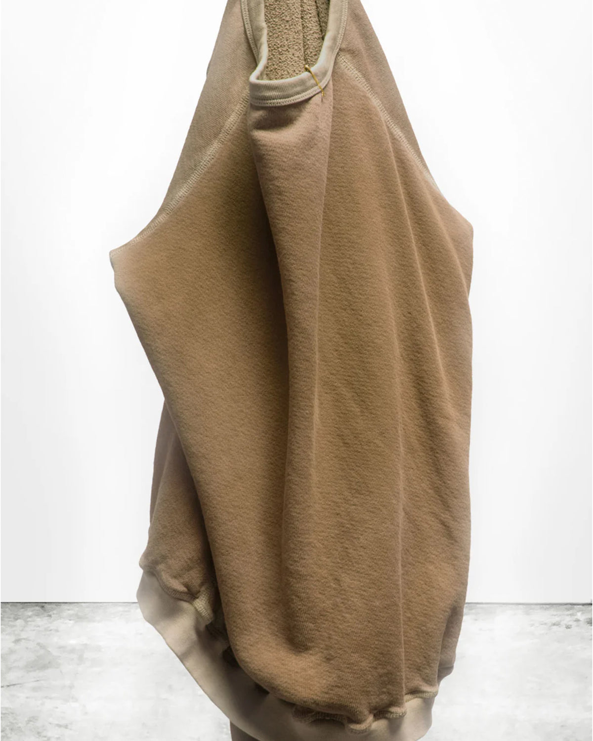 A tan, oversized Lucky Rabbits Sweatshirt by Free City (sparrow, LLC) hangs on a hook against a white wall and concrete floor, featuring lightweight French terry fabric that drapes softly with partially visible sleeves.