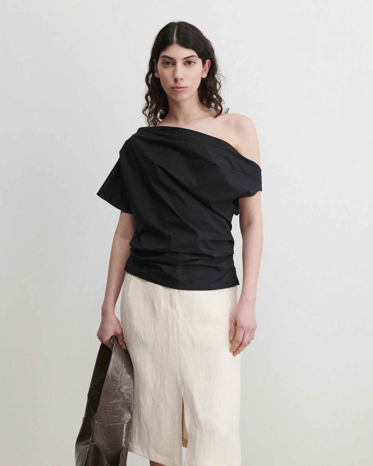 A person with long, dark, wavy hair wears the Rachel Comey Mata Top in black and a cream organic cotton skirt with a front slit, holding a large brown bag against a plain light background.