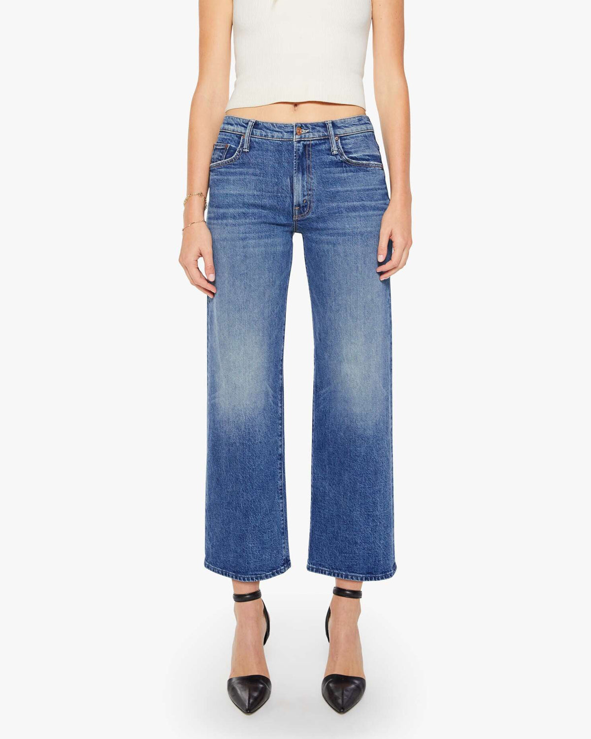 A person wearing Mother’s The Mid Rise Maven Flood jeans in blue, a white sleeveless top, black pointed-toe heels, and a gold bracelet stands against a plain white background.