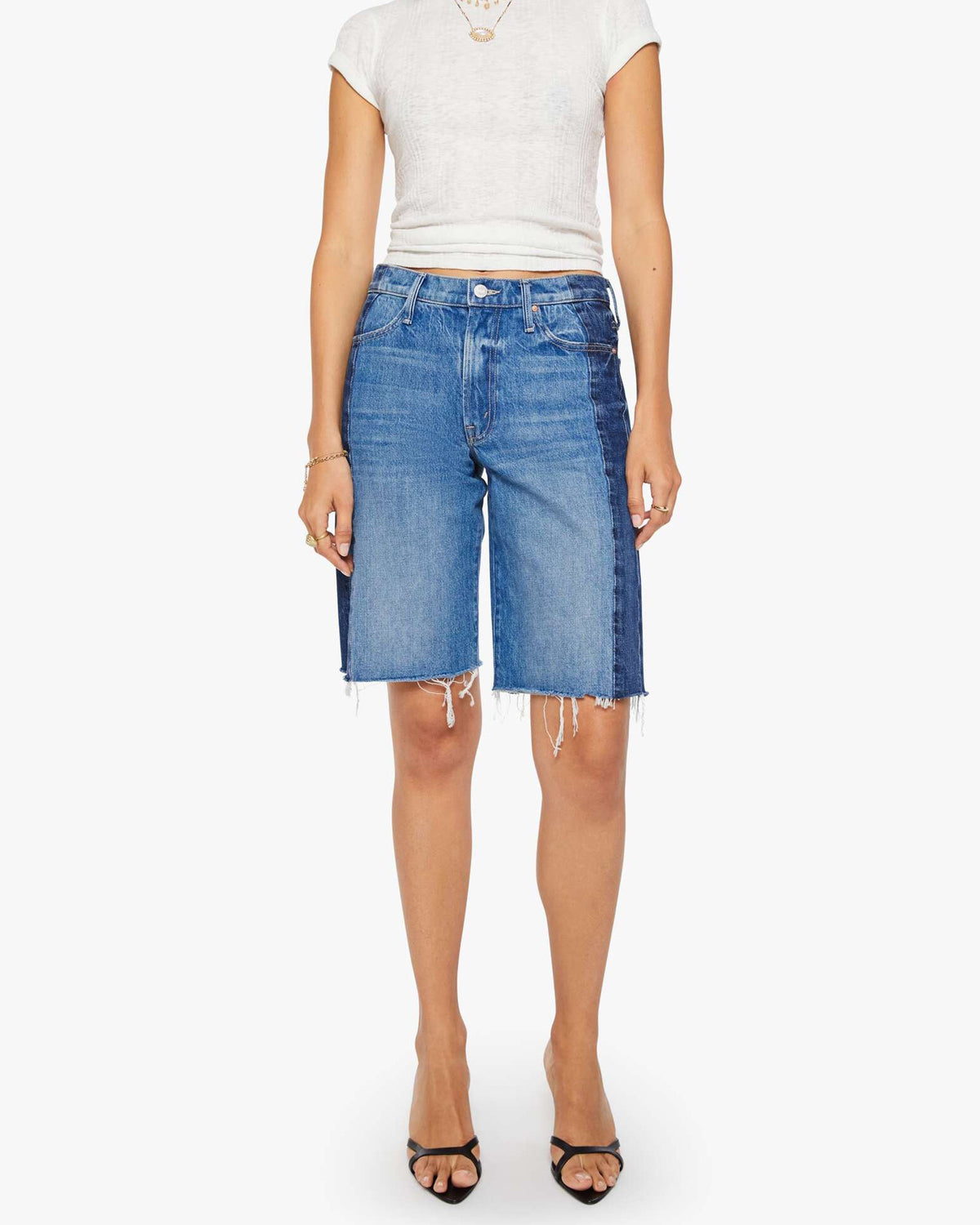 A woman wears a white short-sleeve top and Mother Mid Rise Maven Bermuda Fray denim shorts with a dark side panel, paired with black open-toe heels, standing against a plain white background.