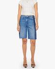 A woman wears a white short-sleeve top and Mother Mid Rise Maven Bermuda Fray denim shorts with a dark side panel, paired with black open-toe heels, standing against a plain white background.