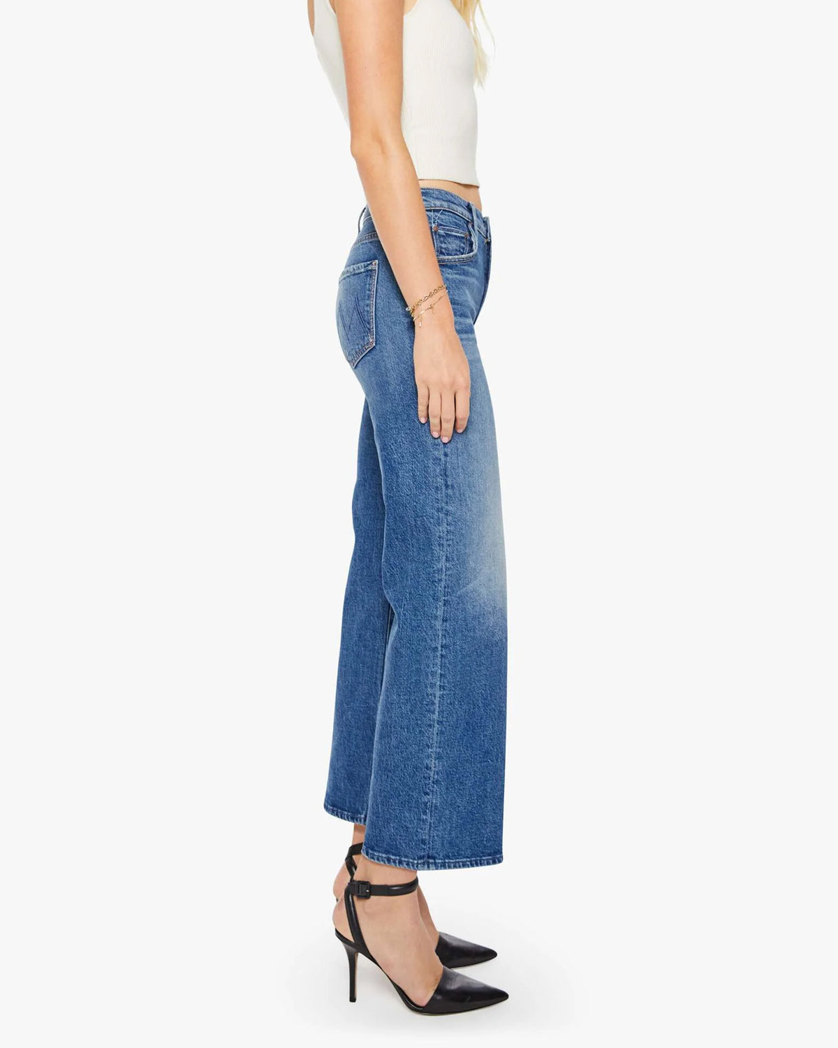 A woman in profile wears Mother’s The Mid Rise Maven Flood—mid-rise, wide-leg blue stretch denim jeans—paired with a white sleeveless top and black heels against a plain white background.