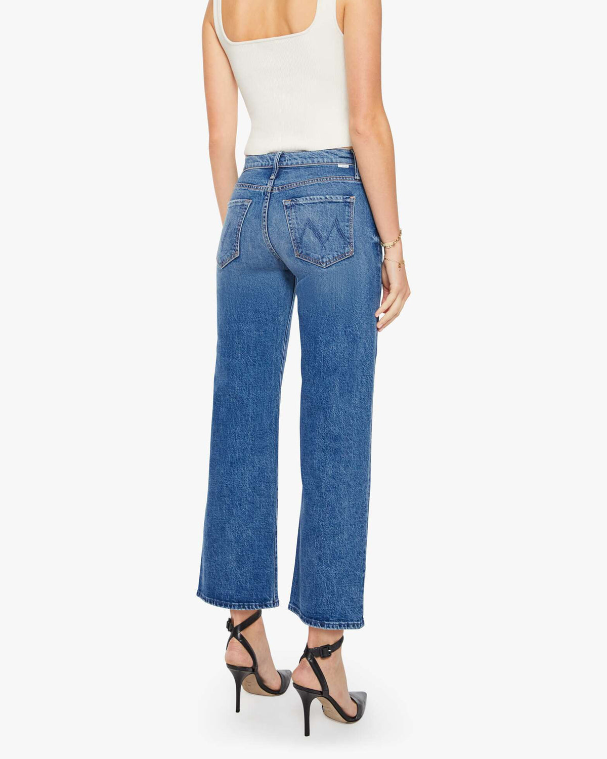 A woman faces away, wearing a white sleeveless top, Mother’s The Mid Rise Maven Flood wide-leg stretch denim jeans with back pockets, and black high-heeled sandals against a plain white background.
