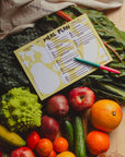 The Faire Meal Planner Notepad, proudly made in the USA, features handwritten days and meals. Shown atop leafy greens with fresh fruits and veggies—like oranges, apples, bananas, cucumbers, and broccoli—inspires healthy planning.