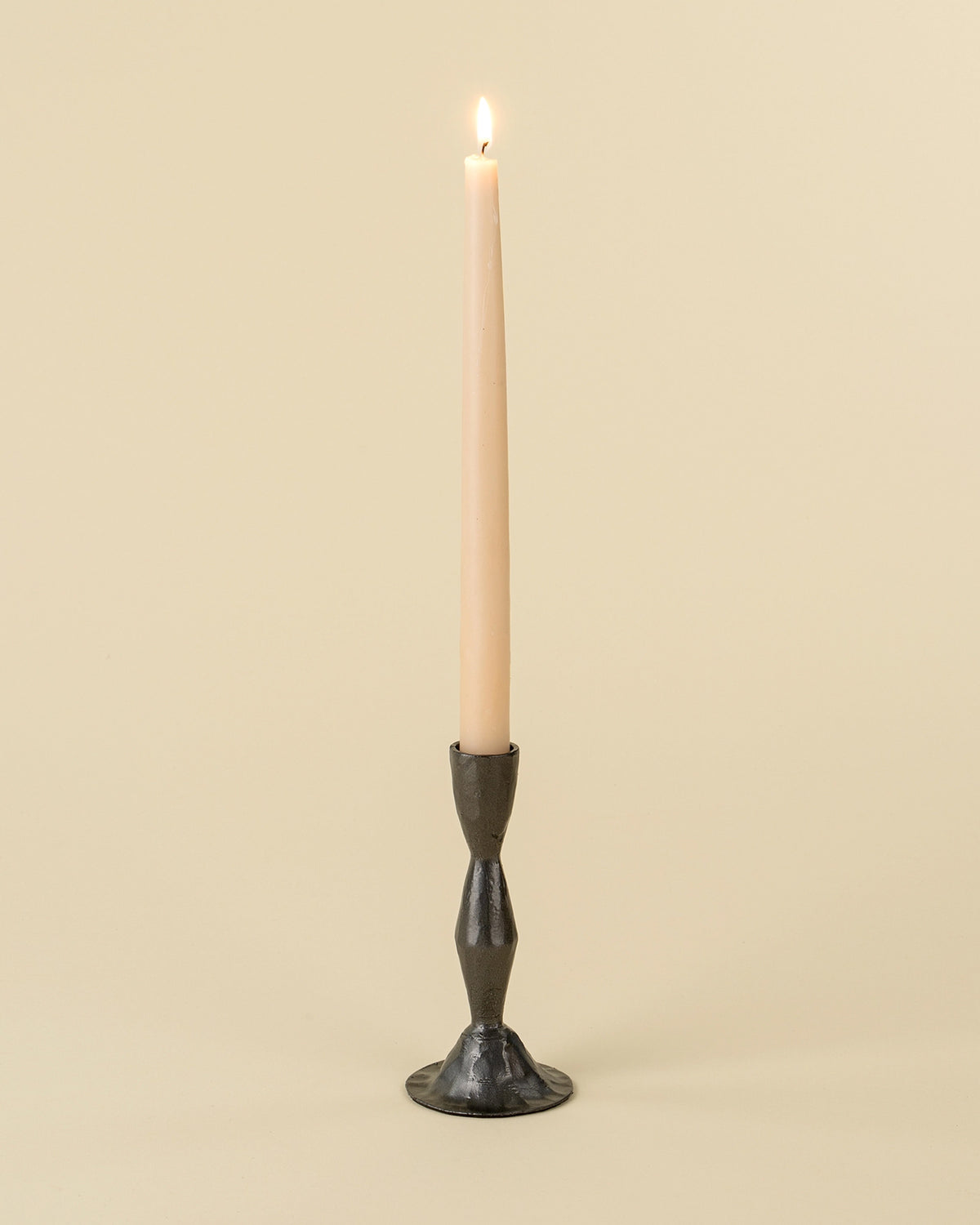 A lit beige taper candle stands in the Indaba 1-2506 Zora Forged Candlestick L, featuring a dark metal finish, set against a light beige background.