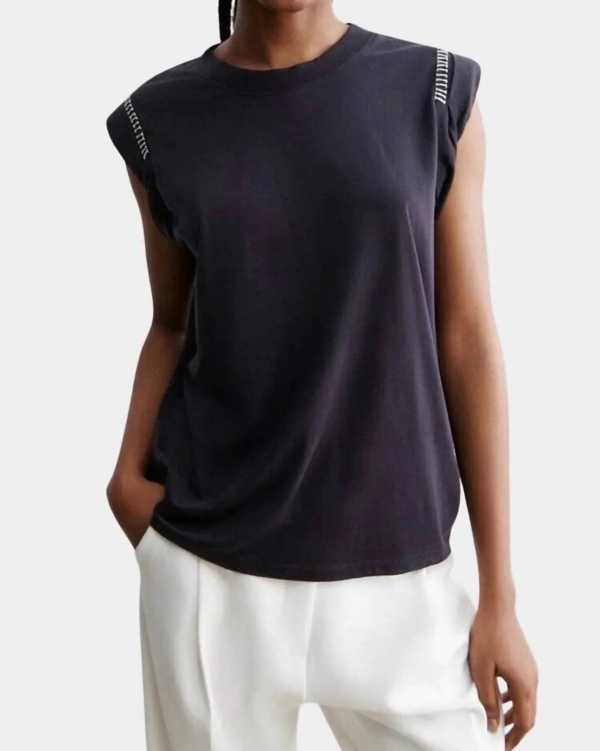 A person wearing the Rachel Comey Miles Tee with white text on the shoulders and loose-fitting white pants stands with one hand in a pocket against a plain background.
