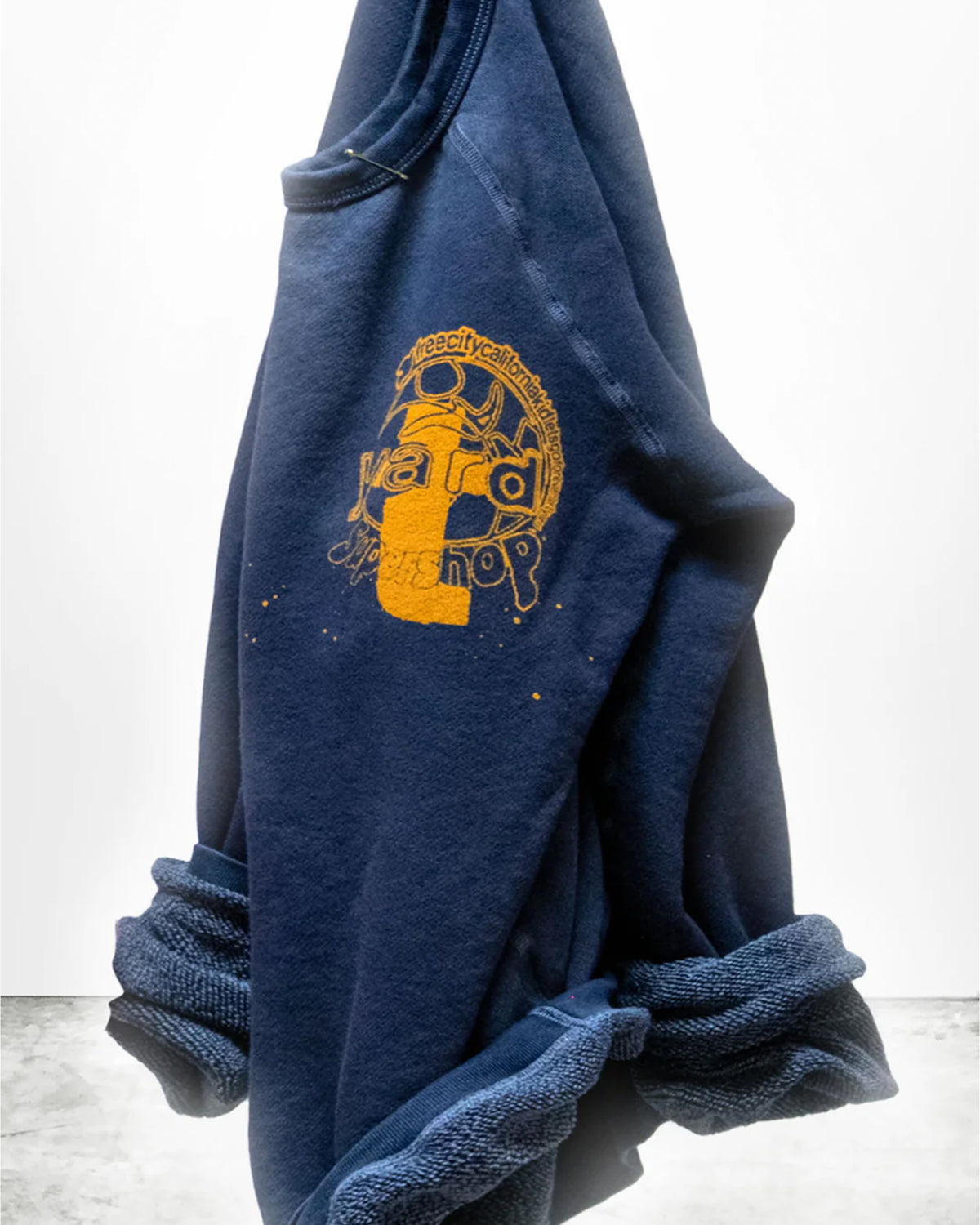 The Artyard SUPERSHOP STAMP Raglan by Free City (sparrow, LLC) is a navy blue unisex sweatshirt with rolled cuffs, made in the USA. It features a large yellow-orange graphic and "thecitycalmbooks yard shop" on the chest.