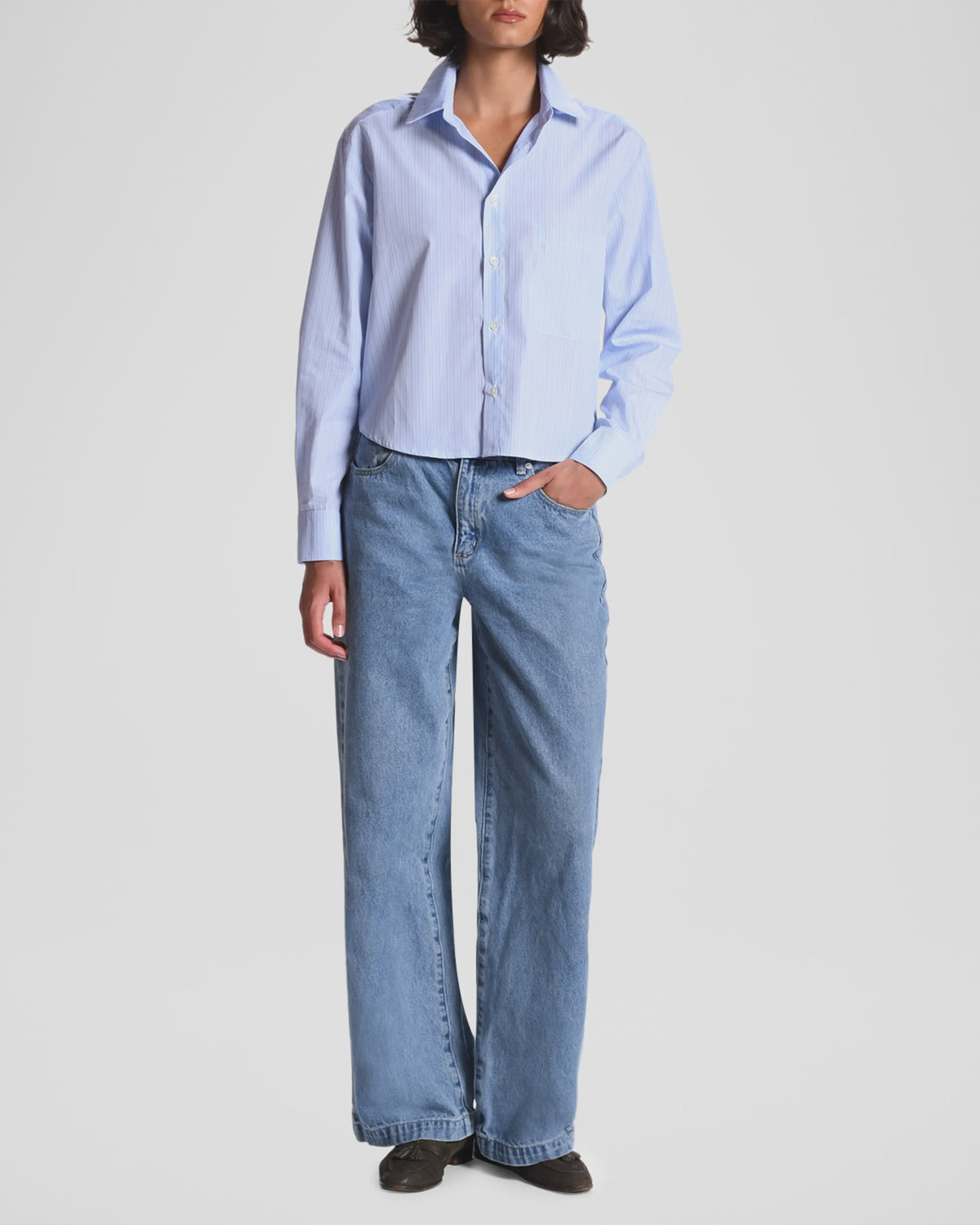 A person, head partially out of frame, stands against a plain background wearing The Belle by Cissa—a relaxed fit light blue button-up shirt—with loose-fitting blue jeans and black shoes, one hand in their pocket.