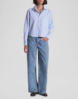 A person, head partially out of frame, stands against a plain background wearing The Belle by Cissa—a relaxed fit light blue button-up shirt—with loose-fitting blue jeans and black shoes, one hand in their pocket.