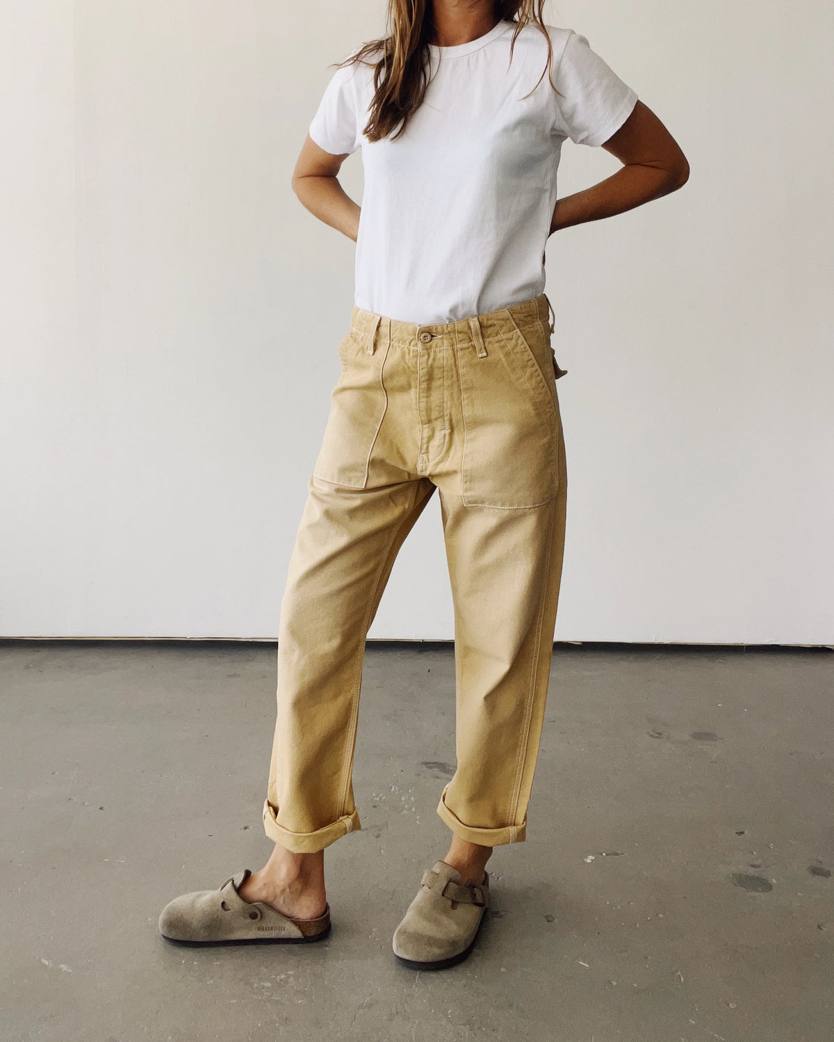 A person stands indoors against a plain wall, wearing a white t-shirt, Hey Gang’s The Officer Pant in cotton ripstop with rolled cuffs, and light brown slip-on shoes. Their hands are on their hips, and their face is out of the frame.