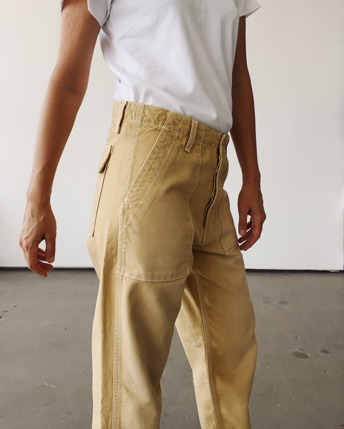 A person stands indoors against a plain white wall, arms at their sides, wearing a white t-shirt and Hey Gang’s The Officer Pant—100% Cotton Ripstop, high-waisted tan work pants with a relaxed leg.