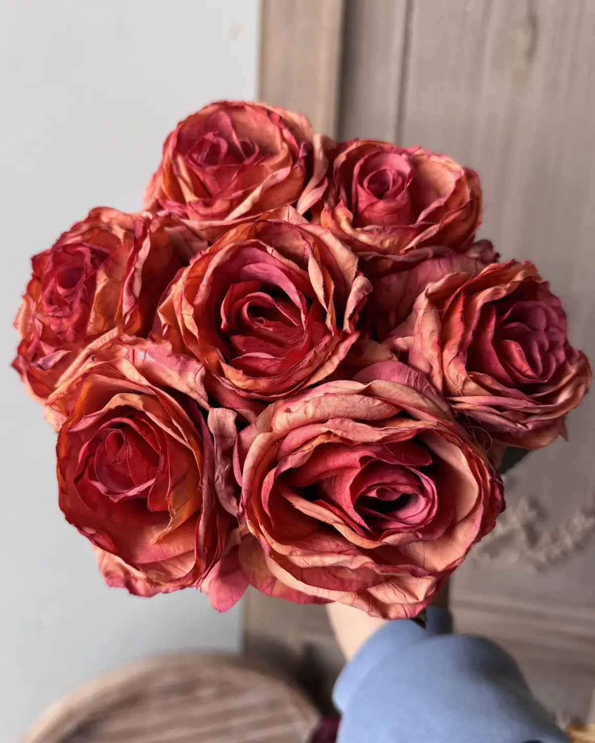 A close-up of a hand holding the Faire 17'' Faux Vintage Rose Bouquet, featuring red and orange blooms with dried, curled petals, set against a blurred wooden background.
