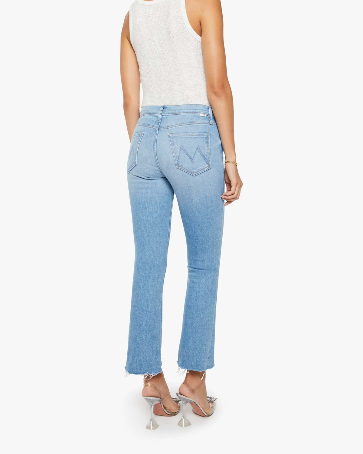 A woman wears Mother’s The Outsider Flood Fray light blue stretch denim bootcut jeans with raw hems, a white sleeveless top, and silver high-heeled sandals, shown from the back against a plain white background.